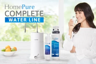 A woman joyfully sipping safe water from a glass, with a full array of Hompure products displayed on the table nearby.