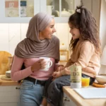 A mother stands while her daughter sits on the kitchen table with a glass of juice next to her. The mother holds a cup, and an EDGE3 multivitamin product is visible on the table