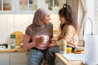 A mother stands while her daughter sits on the kitchen table with a glass of juice next to her. The mother holds a cup, and an EDGE3 multivitamin product is visible on the table