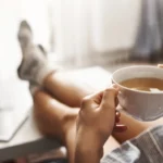 man sitting with legs on table and a cup of coffee in hand