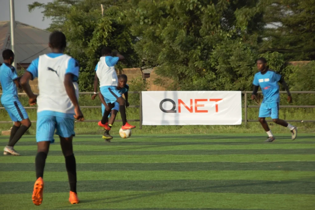 QNET Confederation of african football kids playing on football field with QNET logo behind them. 