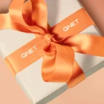 Box with an orange ribbon with QNET branding around it, showing how QNET products are a great way to gift the good life this holiday season