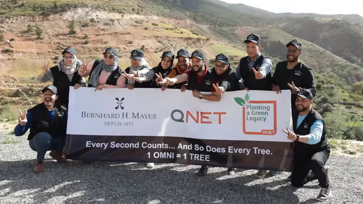 Smiling representatives doing a peace sign as they pose with a banner bearing QNET, Bernhard H Mayer and Green Legacy branding to commemorate having planted a tree for every OMNI watch sold on 7 March 2024 in Amizmiz, Morocco. 