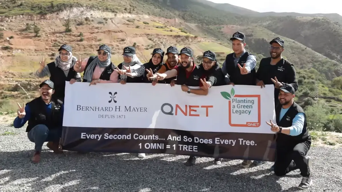 Championing Sustainability And Building A Green Legacy With Qnet 10 Smiling representatives doing a peace sign as they pose with a banner bearing QNET, Bernhard H Mayer and Green Legacy branding to commemorate having planted a tree for every OMNI watch sold on 7 March 2024 in Amizmiz, Morocco.