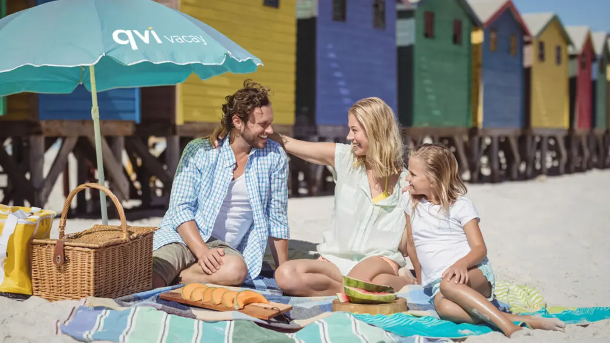 Unlock Your Next Adventure With The Qvi Vacay App! 2 Caucasian family of three sitting on beach towels and enjoying fruit slices under a QVI Vacay umbrella
